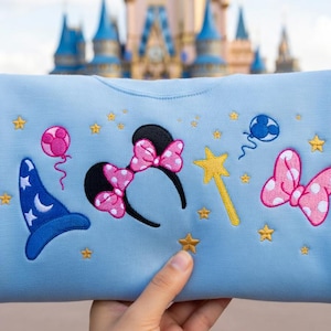 May include: A light blue sweatshirt featuring embroidered Disney-themed designs. The design includes a blue wizard hat, Minnie Mouse ears with a pink bow, a yellow wand, pink bows, and gold stars. The background shows a castle.