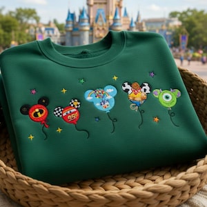 Puede incluir: Una sudadera verde oscuro con globos de personajes de dibujos animados bordados. Los globos representan a Mickey Mouse, un coche de carreras, un globo aerost&aacute;tico, un vaquero y un monstruo verde. La sudadera se muestra en una cesta tejida.