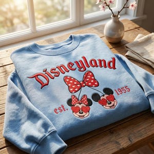 Puede incluir: Sudadera de Disneylandia azul claro con texto rojo bordado y gr&aacute;ficos de Minnie Mouse. El dise&ntilde;o incluye un gran lazo con corazones blancos, dos caras de Minnie Mouse con gafas de sol en forma de coraz&oacute;n y el texto "est. 1955".