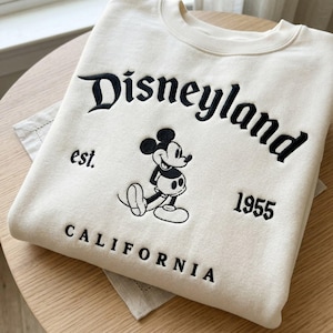 Op de afbeelding: Cr&egrave;mekleurige sweatshirt met zwart geborduurde tekst en een Mickey Mouse-afbeelding. De tekst luidt "Disneyland est. 1955 CALIFORNIA". De sweatshirt ligt op een houten oppervlak.