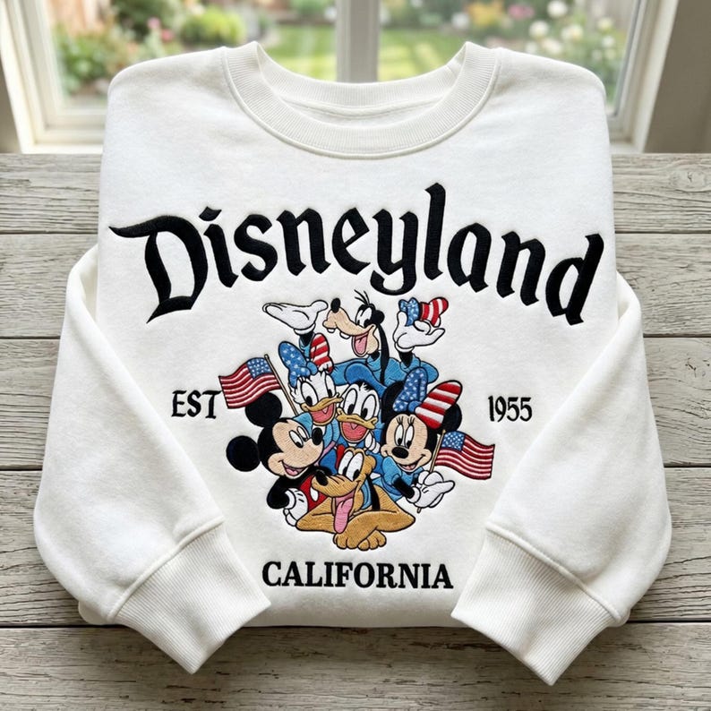 May include: White crewneck sweatshirt with the word "Disneyland" in black script. Features a colorful graphic of Mickey Mouse, Minnie Mouse, Donald Duck, Goofy, and Pluto with American flags. The text "EST 1955 CALIFORNIA" is also included.
