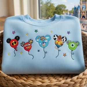 Puede incluir: Sudadera azul claro con globos de personajes de dibujos animados bordados. Los globos presentan orejas de Mickey Mouse y varios dise&ntilde;os de personajes, incluido un globo rojo de Los Incre&iacute;bles, un globo a cuadros con el n&uacute;mero 95 y un globo verde monstruo. La sudadera se muestra en una cesta tejida.