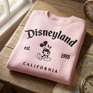 Op de afbeelding: Een lichtroze sweatshirt met het woord "Disneyland" in zwart script. Daaronder een afbeelding van Mickey Mouse, de tekst "est. 1955" en "CALIFORNIA". De sweatshirt is op een houten oppervlak gevouwen.