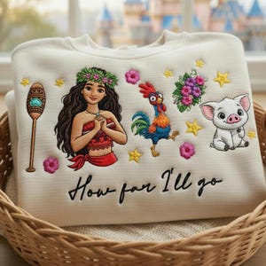 Może przedstawiać: Kremowa bluza z haftowanymi postaciami i napisem "How far I'll go". Projekt zawiera kobietę, koguta, świnię oraz kwiatowe i gwiazdkowe akcenty. Bluza jest prezentowana w wiklinowym koszu.
