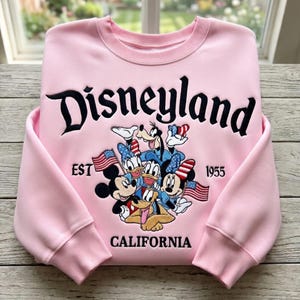 May include: Pink Disneyland sweatshirt featuring Mickey Mouse, Donald Duck, Goofy, and other Disney characters. The sweatshirt has the word "Disneyland" in black script, with "EST 1955 CALIFORNIA" below the characters.