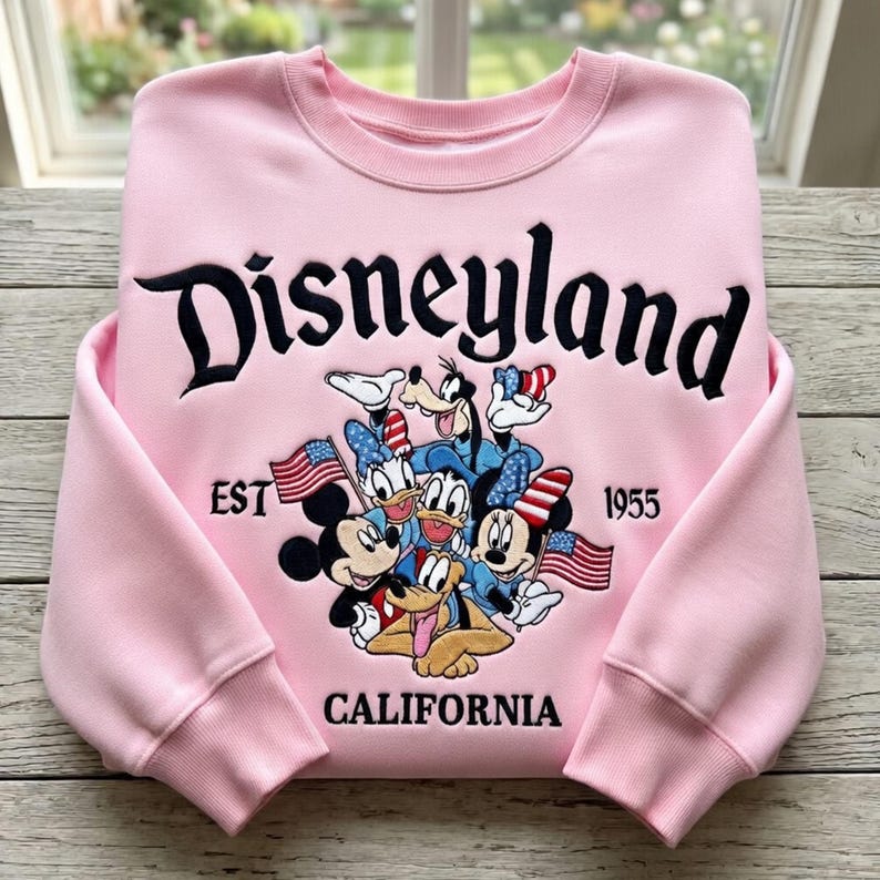 May include: Pink Disneyland sweatshirt featuring Mickey Mouse, Donald Duck, Goofy, and other Disney characters. The sweatshirt has the word "Disneyland" in black script, with "EST 1955 CALIFORNIA" below the characters.