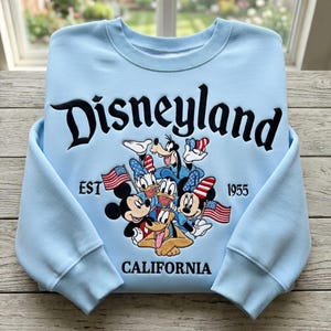 May include: Light blue Disneyland sweatshirt featuring Mickey Mouse, Minnie Mouse, Donald Duck, Daisy Duck, Goofy, and Pluto. The word "Disneyland" is embroidered in black, along with "EST 1955" and "CALIFORNIA". The characters are holding American flags.