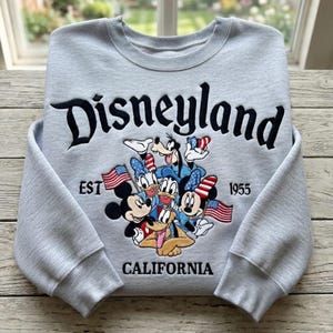 May include: A light gray Disneyland sweatshirt featuring Mickey Mouse, Minnie Mouse, Donald Duck, Daisy Duck, Goofy, and Pluto. The word "Disneyland" is embroidered in black script above the characters, with "EST 1955 CALIFORNIA" below.