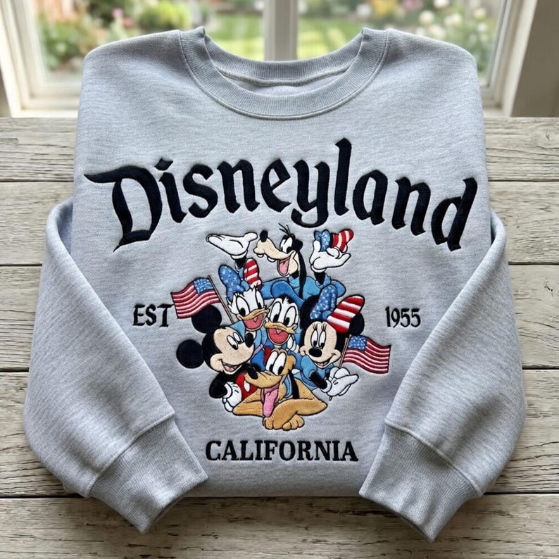 May include: A light gray Disneyland sweatshirt featuring Mickey Mouse, Minnie Mouse, Donald Duck, Daisy Duck, Goofy, and Pluto. The word "Disneyland" is embroidered in black script above the characters, with "EST 1955 CALIFORNIA" below.