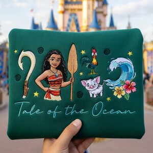 Pu&ograve; includere: Un astuccio verde con immagini ricamate di una donna polinesiana, un amo, una pagaia, un gallo, un maiale, un'onda e fiori. Il testo "Tale of the Ocean" &egrave; ricamato sotto. L'astuccio &egrave; tenuto in mano.