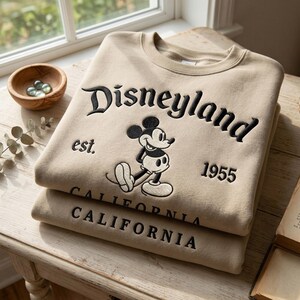 Op de afbeelding: Twee beige sweatshirts liggen op een houten tafel. Het bovenste sweatshirt heeft het woord "Disneyland" in zwarte letters, een Mickey Mouse-afbeelding en de tekst "est. 1955 CALIFORNIA".