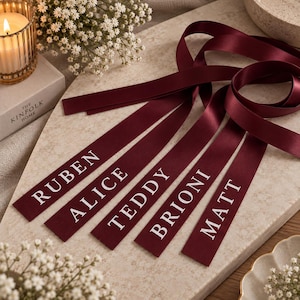 May include: Five long, deep burgundy satin ribbons, each featuring a white printed name. The names displayed on the ribbons are RUBEN, ALICE, TEDDY, BRIONI, and MATT. The ribbons are arranged in a fan-like pattern with smooth, reflective surfaces.