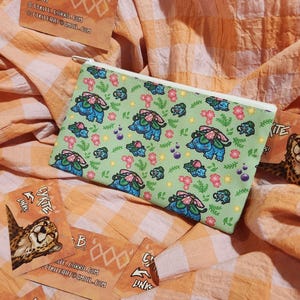 Peut inclure: Une pochette zippée vert clair avec un motif répété de personnages de dessins animés bleus et roses et de fleurs. La pochette est rectangulaire et possède une fermeture éclair blanche. Plusieurs cartes de visite sont éparpillées autour de la pochette.