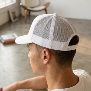 Puede incluir: Gorra de camionero blanca con parte trasera de malla y visera curva. La gorra tiene un cierre a presi&oacute;n ajustable. La persona lleva una camiseta blanca.