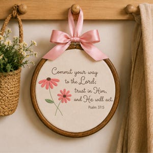 Può includere: Un telaio da ricamo circolare in legno contenente tessuto color avorio con fiori rosa ricamati e il testo: Commit your way to the Lord; trust in Him, and He will act. Psalm 37:5. Un nastro di raso rosa è legato a fiocco in alto.
