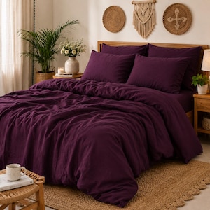 Puede incluir: Un juego de cama de color morado intenso que incluye una funda nórdica y dos fundas de almohada a juego. La tela tiene una apariencia texturizada y ligeramente arrugada. La ropa de cama se dispone sobre una cama con cabecero de madera.