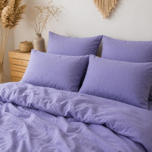 May include: A bedding set featuring a duvet cover and four rectangular pillowcases in a solid light purple color. The fabric has a visible textured weave, and the items are arranged neatly on a bed to showcase the soft, wrinkled appearance.