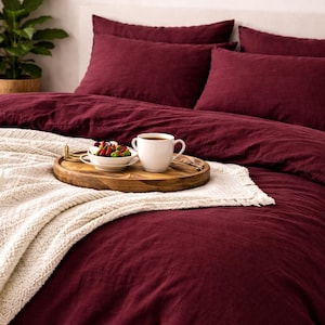 Può includere: Un set copripiumino color bordeaux intenso con federe abbinate, caratterizzato da una coperta color crema testurizzata drappeggiata sul letto. Un vassoio rotondo in legno sul letto contiene una tazza in ceramica bianca e una ciotola di bacche.
