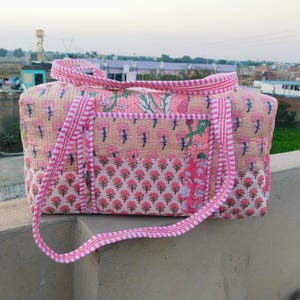 May include: A quilted tote bag with a floral pattern in shades of pink, beige, and green. The bag has a rectangular shape with a top handle and a striped pink trim. The floral design includes stylized flowers and leaves.