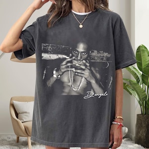 Könnte beinhalten: Dunkelgraues T-Shirt mit einem Schwarz-Weiß-Bild einer Person mit Zigarette und Schmuck. Der Name "D'angelo" steht in Kursivschrift am unteren Rand. Das Shirt hat kurze Ärmel und einen Rundhalsausschnitt.