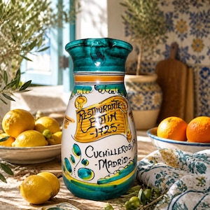 Pode incluir: Um jarro de cerâmica com base branca, borda e base em tom verde-azulado, decorado com detalhes amarelos e verdes. A frente apresenta uma faixa amarela com o texto "Restaurante Botin - 1725" e "Cuchilleros, 17 - Madrid" em preto.