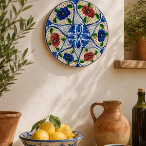Puede incluir: Un plato de pared de cerámica redondo con un patrón floral pintado a mano con flores azules y rojas, hojas verdes y puntos amarillos sobre fondo blanco, dispuesto en un diseño simétrico en forma de estrella con detalles de rejilla azul.