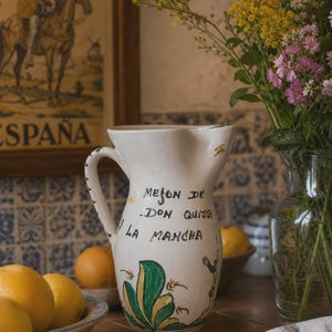 Può includere: Una brocca in ceramica bianca con manico e motivi floreali dipinti a mano in verde e giallo alla base. La parte anteriore mostra il testo nero Meson de Don Quijote La Mancha. La brocca presenta un beccuccio curvo e una finitura liscia.