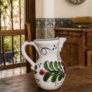 Pode incluir: Um jarro de cerâmica branca com um design floral pintado à mão, apresentando folhas verdes, botões de flores vermelhos e pequenos pontos azuis. O jarro possui uma asa curva, uma borda alargada com bico e um acabamento liso e brilhante.