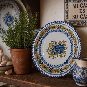 Op de afbeelding: Een rond keramieken bord met een handgeschilderd centraal blauw bloemmotief, omringd door groene en gele bladpatronen. De rand is versierd met een blauw-witte schulprand. Het bord staat op een houten plank.