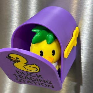 May include: A purple mailbox-shaped magnet with a yellow duck figure inside. The mailbox has a yellow flag and the words "Duck Trading Station" on the door. The duck has a pineapple-like green top.