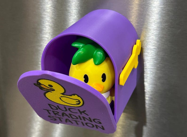 May include: A purple mailbox-shaped magnet with a yellow duck figure inside. The mailbox has a yellow flag and the words "Duck Trading Station" on the door. The duck has a pineapple-like green top.
