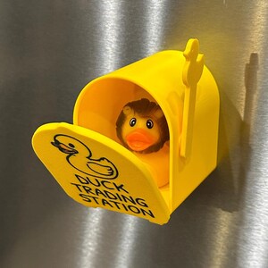 May include: A bright yellow mailbox with a duck-shaped logo and the words "DUCK TRADING STATION" printed on the front. Inside the mailbox is a rubber duck with a brown mane and orange beak. The mailbox is on a metallic surface.