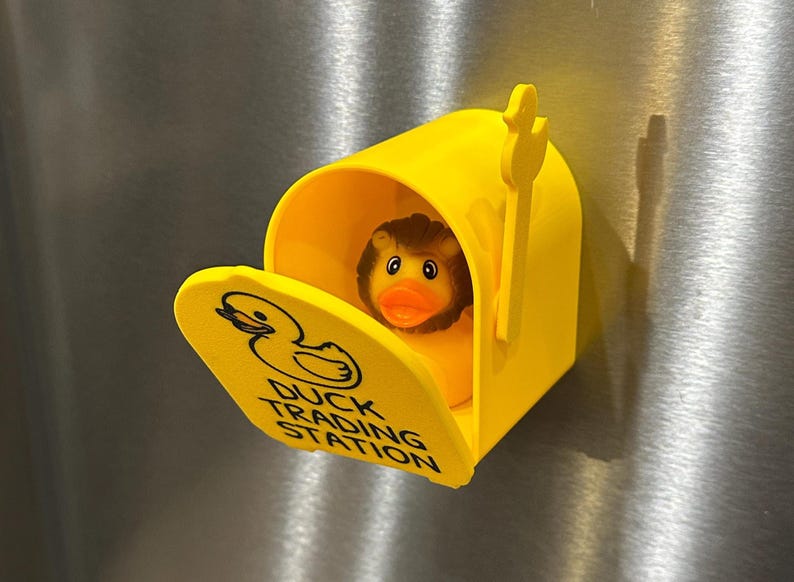 May include: A bright yellow mailbox with a duck-shaped logo and the words "DUCK TRADING STATION" printed on the front. Inside the mailbox is a rubber duck with a brown mane and orange beak. The mailbox is on a metallic surface.