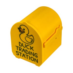 May include: A bright yellow, barrel-shaped container with a duck graphic and the words "DUCK TRADING STATION" in black. The container has a curved top and a small opening on the side.