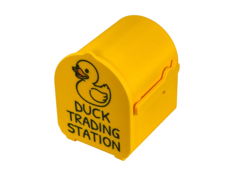 May include: A bright yellow, barrel-shaped container with a duck graphic and the words "DUCK TRADING STATION" in black. The container has a curved top and a small opening on the side.