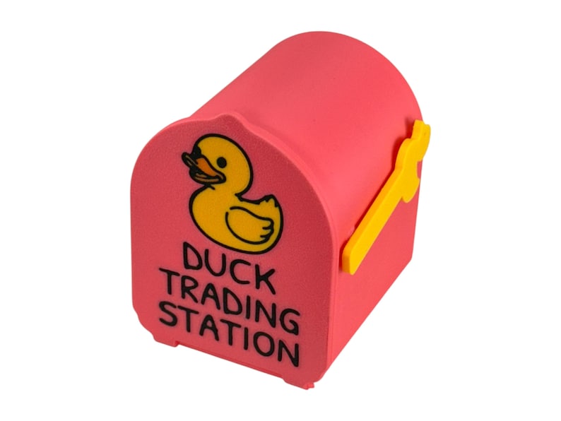 May include: A pink mailbox-shaped container with a yellow duck illustration and the text "DUCK TRADING STATION." The mailbox has a yellow handle and is designed to resemble a traditional mailbox.