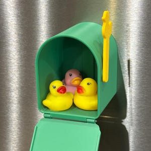 May include: A green mailbox with a yellow flag houses three rubber ducks. Two yellow ducks with red beaks flank a pink duck. The mailbox is mounted on a metallic surface.
