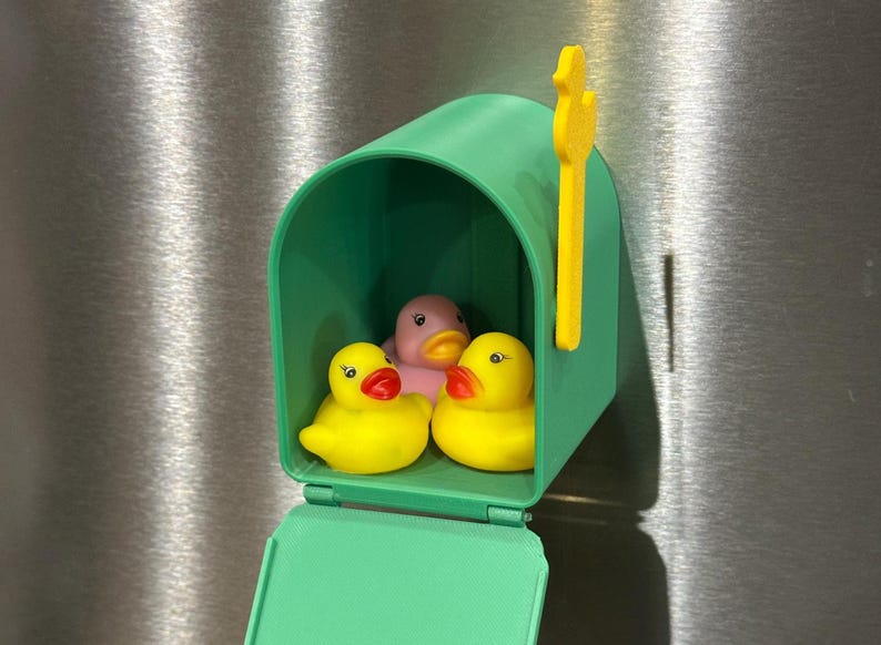 May include: A green mailbox with a yellow flag houses three rubber ducks. Two yellow ducks with red beaks flank a pink duck. The mailbox is mounted on a metallic surface.