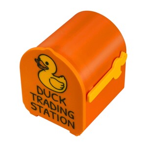 May include: Orange mailbox-shaped container with a yellow duck logo and the words "Duck Trading Station." The container has a yellow handle and is designed to resemble a miniature mailbox.