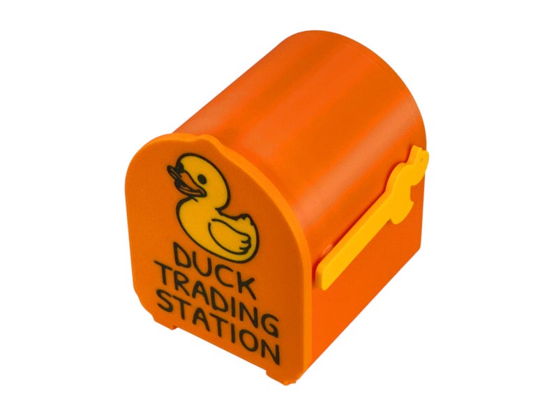 May include: Orange mailbox-shaped container with a yellow duck logo and the words "Duck Trading Station." The container has a yellow handle and is designed to resemble a miniature mailbox.