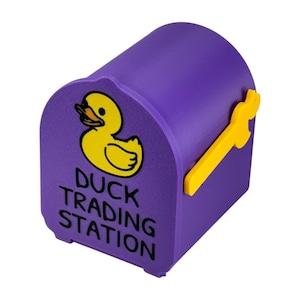 May include: A purple mailbox-shaped container with a yellow duck illustration and the text "DUCK TRADING STATION." The mailbox has a yellow handle and is designed to resemble a miniature mailbox.