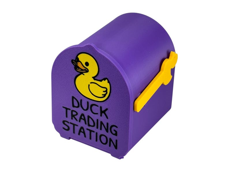 May include: A purple mailbox-shaped container with a yellow duck illustration and the text "DUCK TRADING STATION." The mailbox has a yellow handle and is designed to resemble a miniature mailbox.