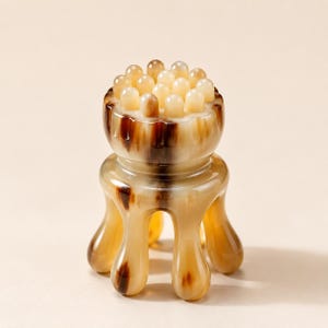 Peut inclure: Un outil de massage manuel en corne polie avec un motif marbré brun et crème. L'objet présente un sommet circulaire doté de multiples picots arrondis pour la pression et quatre pieds incurvés s'étendant vers le bas pour une prise stable.
