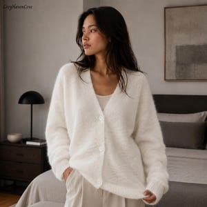 May include: A fuzzy, off-white V-neck cardigan featuring long sleeves, ribbed cuffs, and a front button closure with three matching white buttons. The knit garment has a soft, textured appearance and is worn over a simple, light-colored base layer.