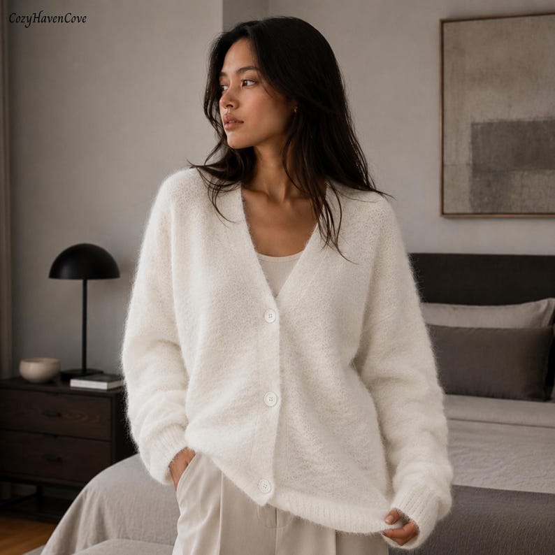 May include: A fuzzy, off-white V-neck cardigan featuring long sleeves, ribbed cuffs, and a front button closure with three matching white buttons. The knit garment has a soft, textured appearance and is worn over a simple, light-colored base layer.