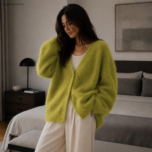 May include: A fuzzy, lime green V-neck cardigan with long sleeves and a single button closure, worn over a plain white tank top and light-colored trousers. The knit fabric has a soft, textured appearance with a visible, slightly loose weave.
