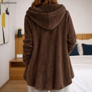 May include: A brown hooded jacket made of a soft, fuzzy material. The jacket has long sleeves and a hood. The person is wearing gray sweatpants. The Cozy Haven Cove logo is in the top left corner.
