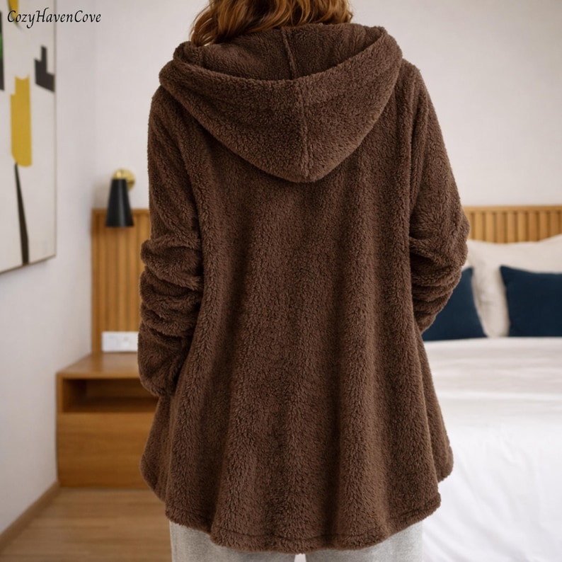 May include: A brown hooded jacket made of a soft, fuzzy material. The jacket has long sleeves and a hood. The person is wearing gray sweatpants. The Cozy Haven Cove logo is in the top left corner.