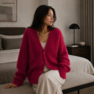 May include: A vibrant magenta V-neck cardigan featuring a fuzzy, textured knit fabric, long sleeves with ribbed cuffs, and a front button closure with three matching buttons, worn over a simple light-colored top and loose-fitting trousers.