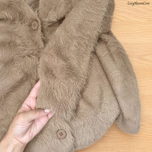 May include: A fuzzy, light brown cardigan with long sleeves and two visible round buttons. The knit fabric has a soft, textured appearance. The text CozyHavenCove is printed in the upper right corner of the image.